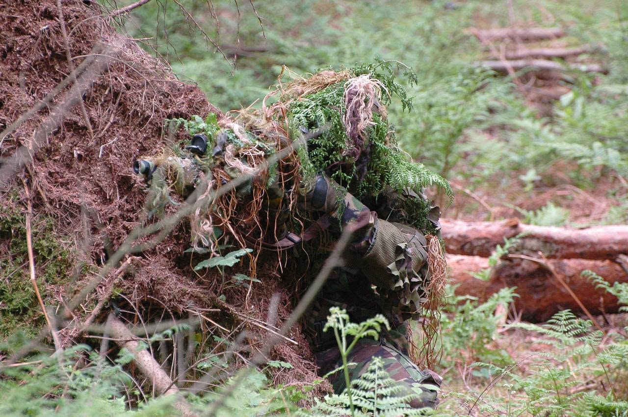 Dale (TheBauer) ghillied up in full woodland concealment, aiming a scoped airsoft sniper rifle