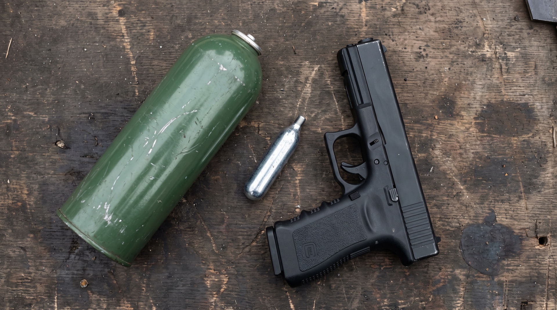 A green gas canister, a CO2 capsule and a gas blowback airsoft pistol arranged on a dark surface
